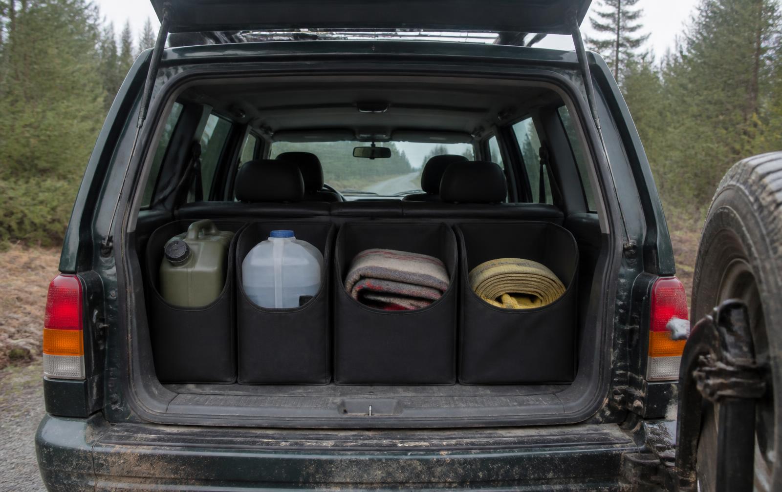 Open SUV hatch on a forest gravel road showing four black nylon compartments holding a fuel can, water jug, blanket and recovery strap